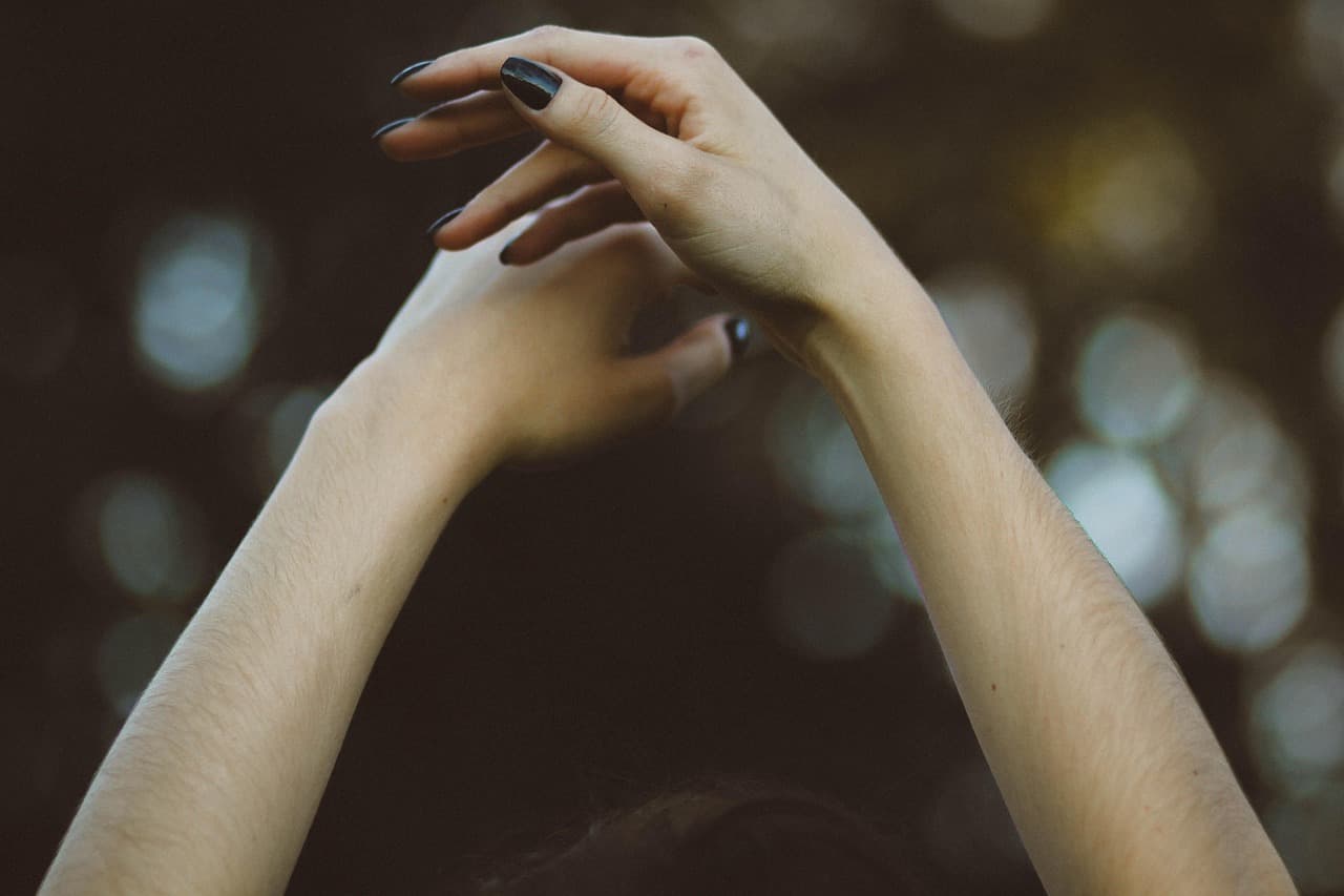 Two raised hands with long, glossy black almond nails against a dark background with soft out-of-focus lights